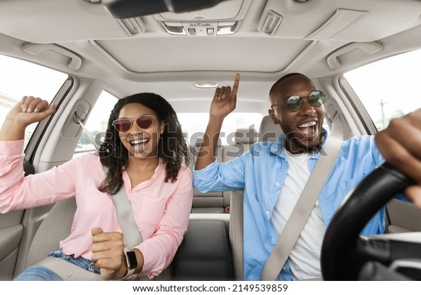 Road Trip Concept. Portrait of excited playful\
African American couple in sunglasses driving luxury car, dancing\
to music singing favorite song, resting on weekend going on\
vacation, windshield view