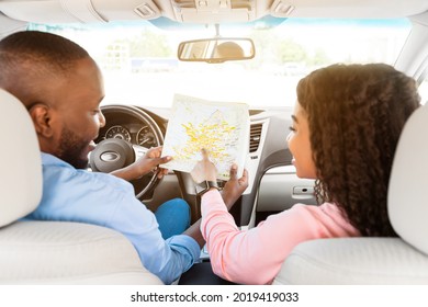 Road Trip Concept. Rear Back View From Passenger's Seat Of Smiling Black Man And Lady Holding And Looking At Paper Map Sitting Inside Car, Choosing Location, Woman Pointing At Destination Point