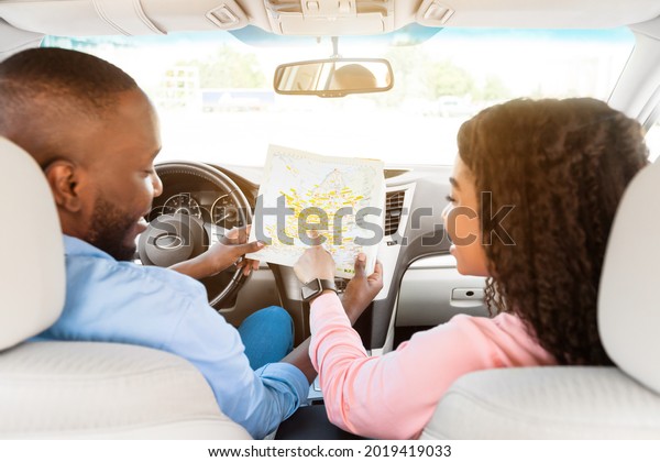 Road Trip Concept. Rear back view from passenger\'s\
seat of smiling black man and lady holding and looking at paper map\
sitting inside car, choosing location, woman pointing at\
destination point