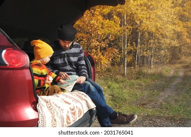 Road Trip In Fall Season, Family Travel, Self-guided Journey. Father And Child Boy Looking On Map And Pointing Route, While Sitting In Car Trunk In Autumn Forest.