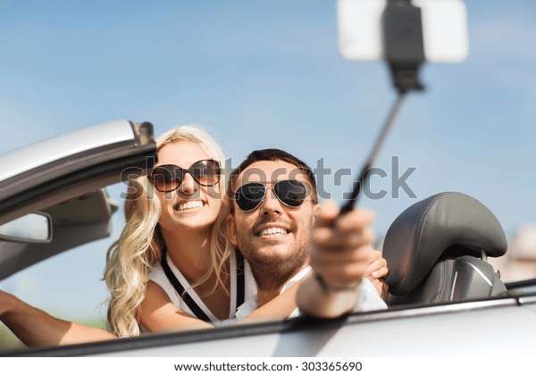 road trip, leisure, couple,
technology and people concept - happy man and woman driving in
cabriolet car and taking picture with smartphone on selfie
stick