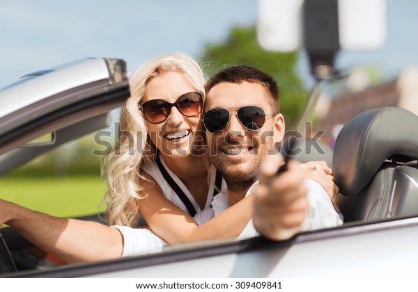 road trip, leisure, couple,
technology and people concept - happy man and woman driving in
cabriolet car and taking picture with smartphone on selfie
stick