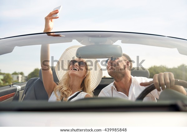 road trip, leisure, couple, technology and\
people concept - happy man and woman driving in cabriolet car and\
taking selfie with\
smartphone