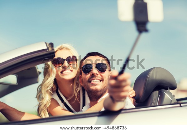 road trip, leisure, couple,\
technology and people concept - happy man and woman driving in\
cabriolet car and taking picture with smartphone on selfie\
stick