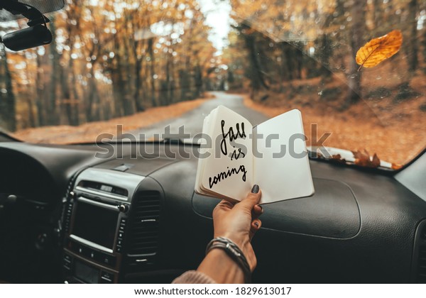 Road trip looking inside of car.\
Woman hand holding notebook with Fall Is Coming text over\
windshield. Freedom travel concept. Autumn weekend to the\
forest.