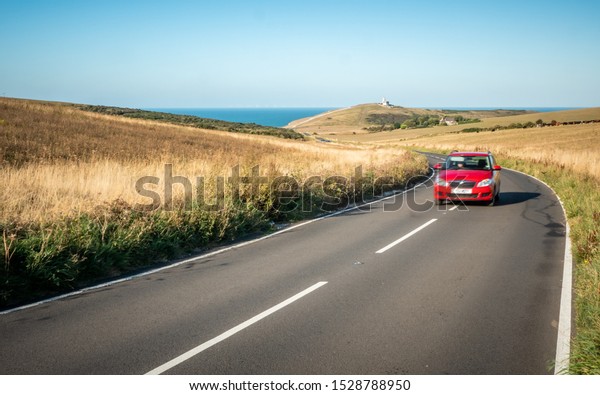 Road Trip; South Downs, England. A car\
travelling a winding road in East Sussex with Belle Tout lighthouse\
and the English Channel in the\
distance.