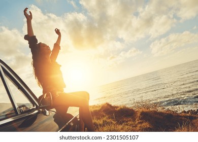 Road trip, sunset and arms raised with a woman at the coast, sitting on her car bonnet during travel for freedom or escape. Nature, flare and water with a young female tourist traveling in summer - Powered by Shutterstock