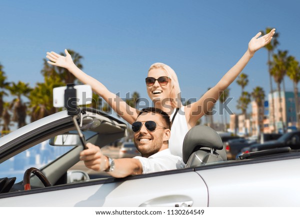 road trip,\
technology and travel concept - happy couple driving in convertible\
car and taking picture by smartphone on selfie stick over venice\
beach background in\
california