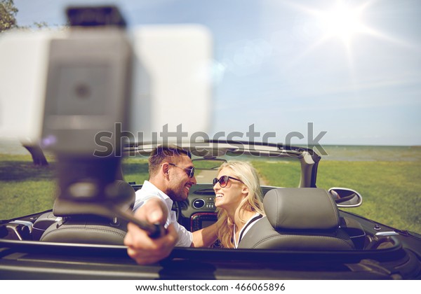 road trip, travel, couple, technology and
people concept - happy man and woman driving in cabriolet car and
taking picture with smartphone on selfie
stick