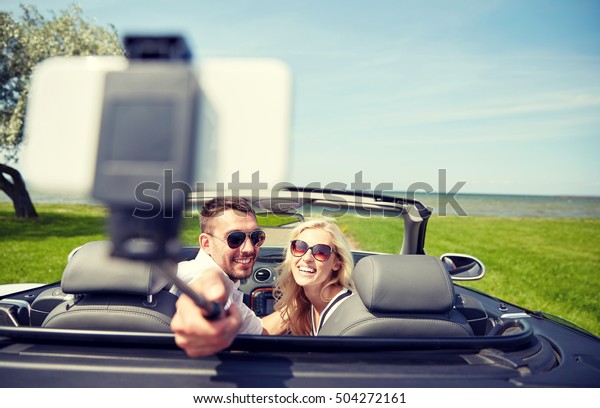 road trip, travel, couple, technology and\
people concept - happy man and woman driving in cabriolet car and\
taking picture with smartphone on selfie\
stick