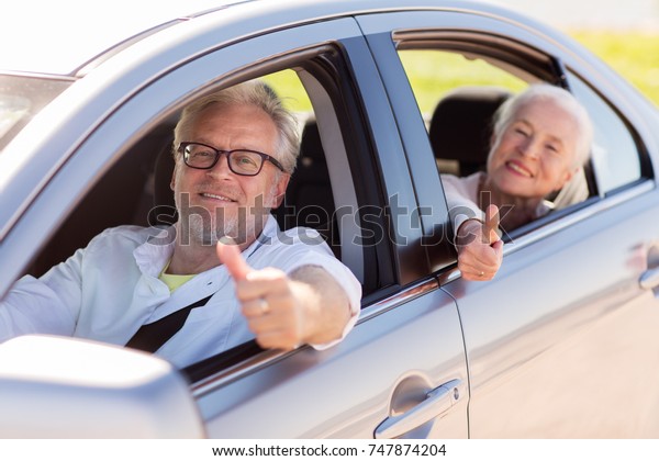 road trip, travel and old\
people concept - happy senior couple driving in car and showing\
thumbs up