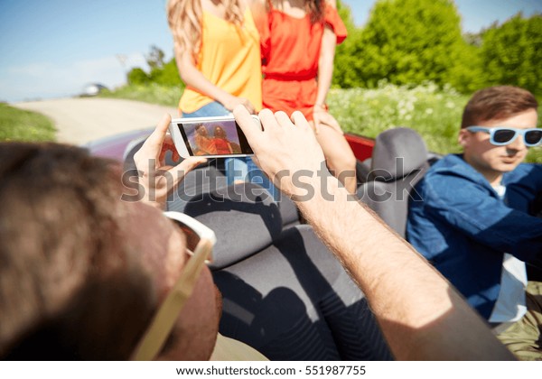 road trip, travel, technology and people concept\
- happy friends driving in cabriolet and photographing by\
smartphone outdoors