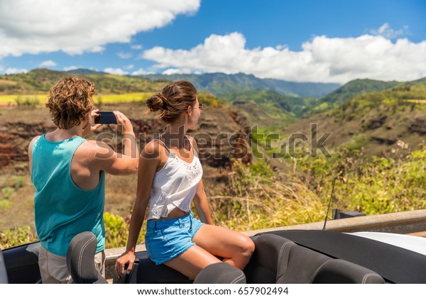 Road trip\
vacation car couple taking pictures of Hawaii nature landscape with\
smartphone camera app. Young people relaxing in convertible car on\
summer holidays. Travel\
lifestyle.