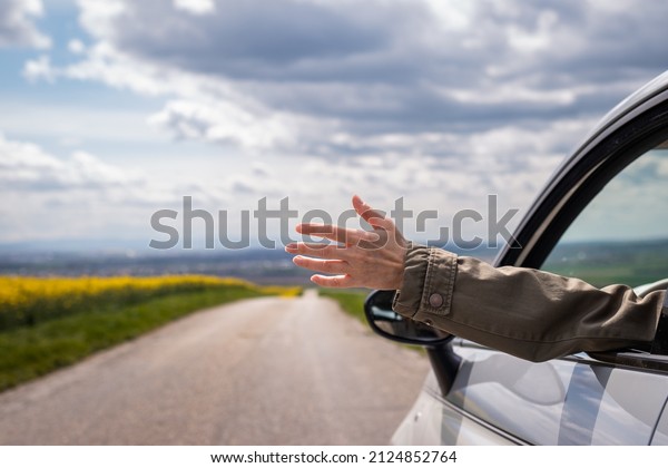 Road trip. Woman waving hand\
of car window during driving at country road. Enjoyment of\
vacation