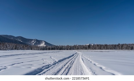 The Road, Trodden In The Snow, Goes Forward And Into The Distance Along A Spacious Valley. Clean Untouched Snow-covered Fields Around. The Taiga Is Visible Ahead. Mountains Against A Clear Blue Sky.