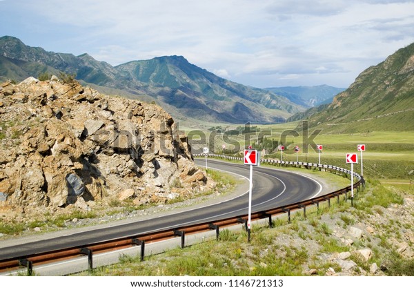 Road turn of the asphalt road with a dividing\
strip, with road markings and red signs of turn. The road is\
surrounded by green hills and mountains. Chuya highway in the\
valley of the river\
Chuya.Altai