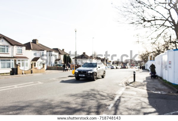Road UK, black car\
driving on the street, residential road, people walking on the side\
walk, blurred.