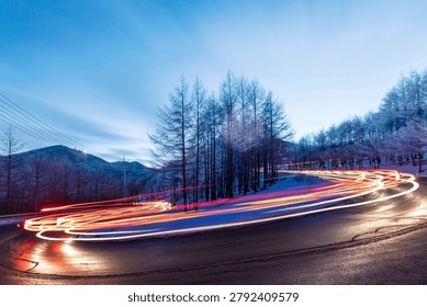 A road winding in a sharp S-shape in a high mountainous region surrounded by lush forests—this is a valuable photograph captured from a high vantage point, showcasing the main view of a motorcycle speeding through the beautiful scenery of the mountain slopes, along with the colorful light trails and dynamism of cars. It is suitable for general content planning, editing, and commercial design.