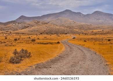 A road winds through a desert with mountains in the background. The road is rocky and dusty, and the landscape is barren and desolate. Scene is one of loneliness and isolation - Powered by Shutterstock