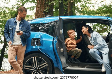 Road in woodland. Mother, father and little son are waiting for electric car to charge outdoors.