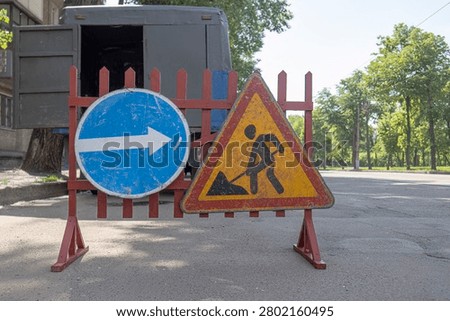 Road Work Triangular Sign and Blue Arrow Detour Sign Mounted on Red Barrier Fence on City Street Context Shot