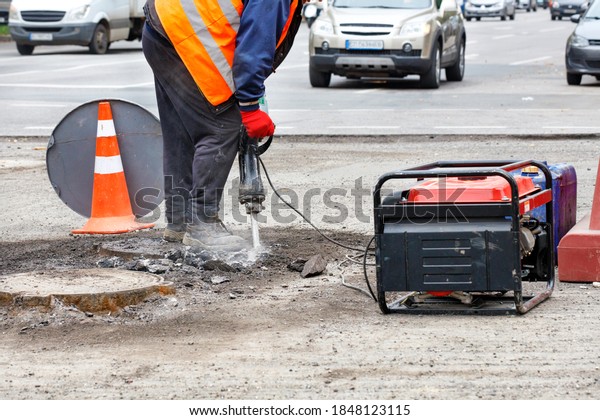 A road worker, dressed in
reflective clothing, repairs a section of road near sewers with an
electric jackhammer, with passing cars in blur as background, copy
space.
