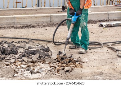 A road worker in green overalls loosens old asphalt with a two-handed pneumatic jackhammer at a road workplace on a summer day. Copy space. - Shutterstock ID 2096414623