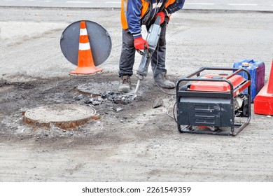 Trabajador de la carretera en un día despejado reparando la carretera de una sección de la carretera con un jackhammer eléctrico y un generador de gasolina cerca de los agujeros de alcantarillado. Copiar espacio.