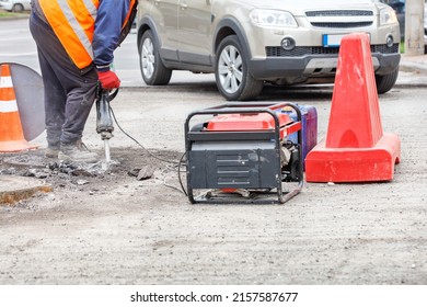 A road worker in a reflective orange vest is repairing a section of road in a fenced area around sewer manholes using an electric jackhammer and a gasoline generator. Copy space. - Shutterstock ID 2157587677