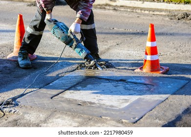 A Road Worker Using A Pneumatic Jackhammer Breaks The Asphalt At The Sewer Hatch. Noisy Drill. Road Construction Works.