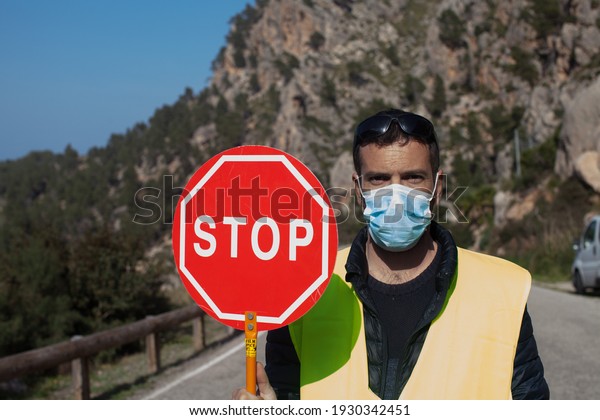 A\
road worker with a walkie talkie and a reflective vest while\
holding a stop sign, the worker wears a surgical\
mask
