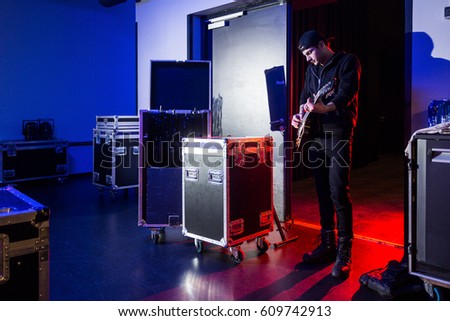 Roadie playing guitar backstage, standing near the stage entrance, surrounded by flight cases, containing equipment.