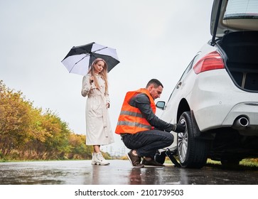 Roadside Assistance Worker Replacing Flat Tire While Beautiful Woman In Trench Coat Holding Umbrella. Male Auto Mechanic Changing Flat Tire On Woman Car The Road. Concept Of Emergency Road Service.