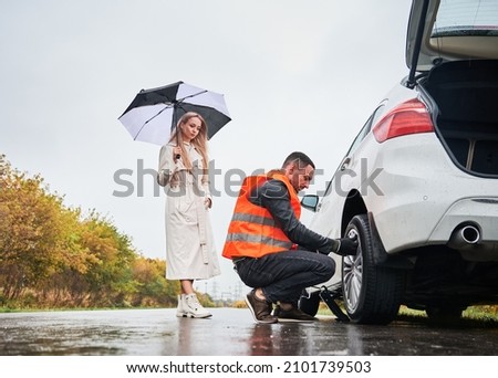 Roadside assistance worker replacing flat tire while beautiful woman in trench coat holding umbrella. Male auto mechanic changing flat tire on woman car the road. Concept of emergency road service.