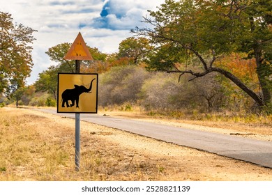 A roadside warning sign featuring an elephant silhouette, alerting travelers to the presence of wildlife on a rural road surrounded by trees and open landscape, under a partly cloudy sky. - Powered by Shutterstock