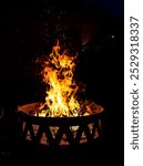 A roaring bonfire illuminates the night sky with dancing flames and sparks. The firepit casts a warm glow on the surrounding area, creating a cozy and inviting atmosphere.