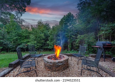 Roaring fire in a stone firepit with wood logs and surrounded by trees at dusk in a backyard