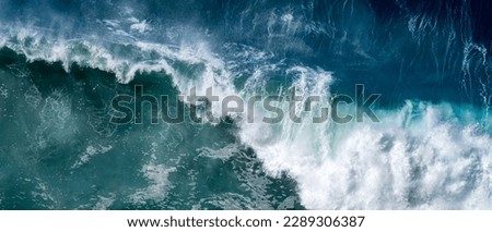 A roaring wave crashes into the deep waters, creating a mesmerizing display of power and beauty, with its frothy white foam contrasting against the deep blue ocean, captured from a high vantage point.