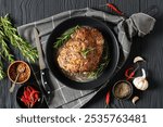 Roast Pork Neck on a pan on black wooden table with dried red pepper, rosemary, garlic, bay leaves, knife, landscape view from above