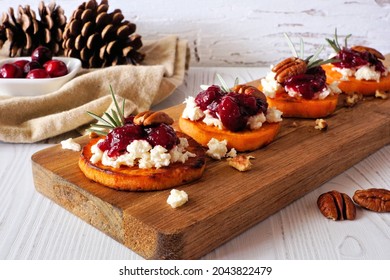Roasted autumn sweet potato crostini appetizers with cheese, cranberries and pecans on a wood platter. Table scene with a white wood background.