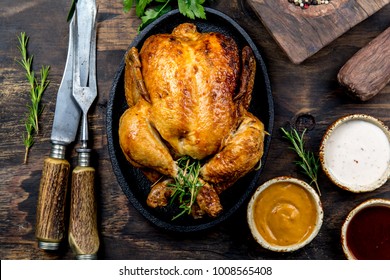 Roasted chicken with rosemary served on black plate with sauces on wooden table, top view