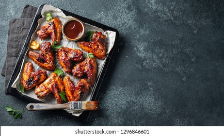 Roasted chicken wings in barbecue sauce with sesame seeds and parsley in a baking tray on a dark table. Top view with copy space. Tasty snack for beer on a dark background. Flat lay.