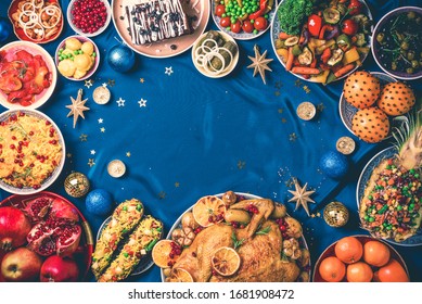 Roasted Christmas turkey with orange slices, cranberries, garlic, festive decoration, candles, tangerine, pomegranate, golden cultery, star glitter sparkles on blue background. Top view, copy space