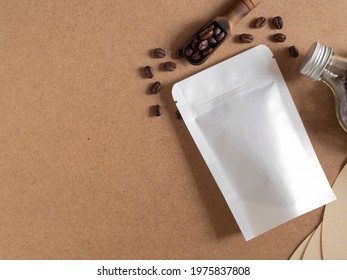 Roasted Coffee Beans In White Paper Bag With Zipper And Wooden Scoop, Glass Bottle On Brwon Background. Space For Your Text, Flat Lay, Top Down.