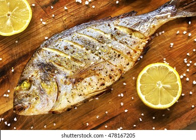 Roasted gilt head bream fish on a wooden table with lemons and coarse grained salt.