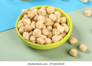Roasted hazelnut kernels in a bright yellow bowl on a colorful blue and green background with scattered nuts, perfect for food and snack photography.