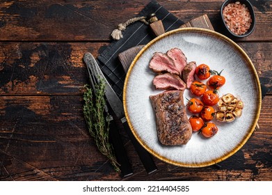 Roasted Lamb tenderloin meat in plate with grilled tomato and garlic, mutton sirloin fillet steak. Dark background. Top view. Copy space. - Shutterstock ID 2214404585
