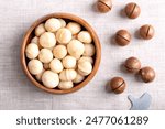Roasted macadamia nuts in a wooden bowl on linen fabric. On the right fruits with sawn nutshells and opener key. Also known as Queensland, bush, maroochi, bauple and Hawaii nuts. Close-up, from above.