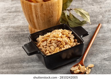 Roasted peanuts fill black square bowl beside wooden spoon holding few nuts. Background features wooden mortar dried leaves. Warm earth tones, soft lighting, rustic food styling