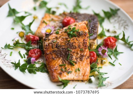 Roasted red salmon trout (steelhead) with tomato and decorative dandelion leaf and edible daisy flower on plate.